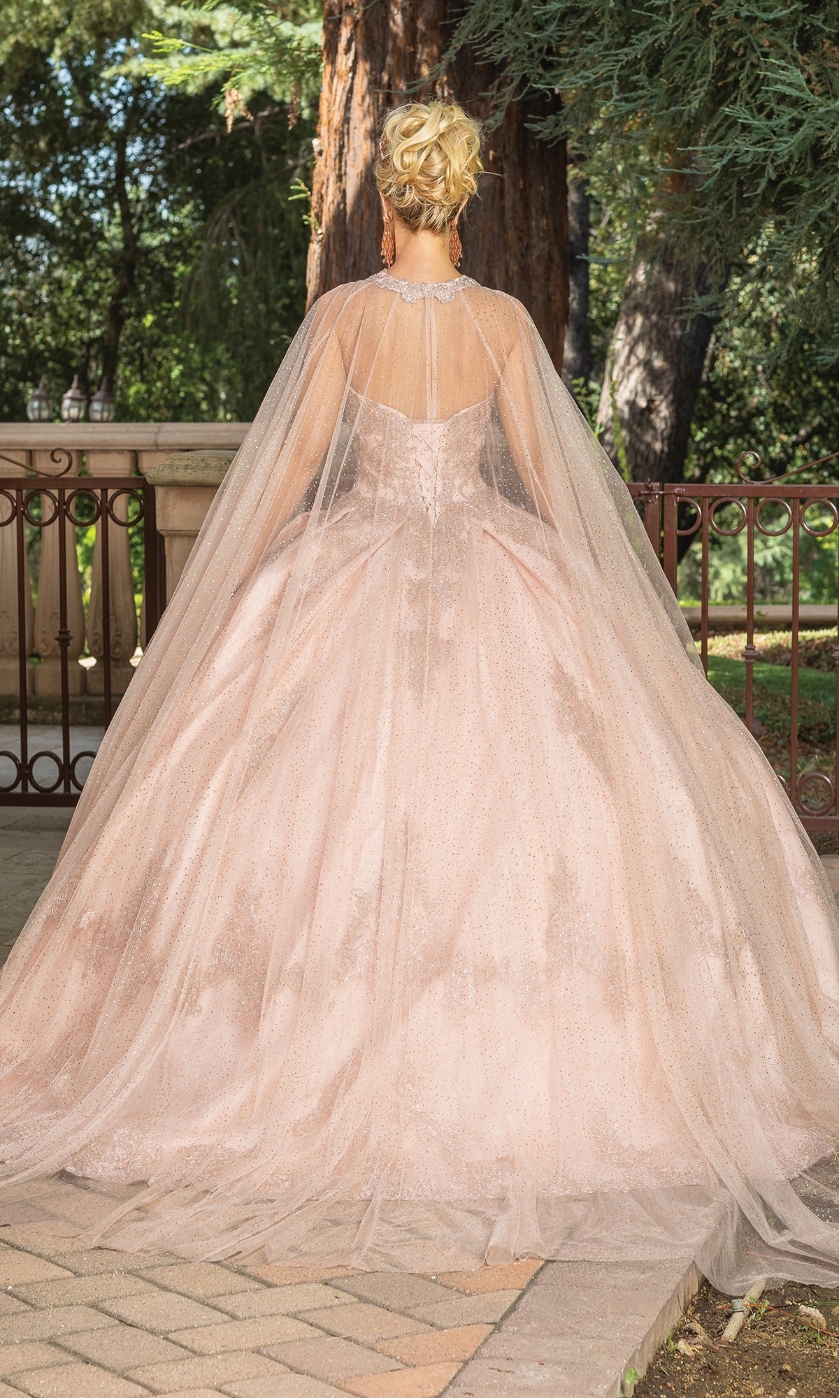 A woman poses outdoors before trees, elegantly showcasing the Quinceanera Dress 1771 by Dancing Queen—a rose gold, light pink a-line ball gown with a sheer cape and strapless sweetheart neckline—seen from the back.